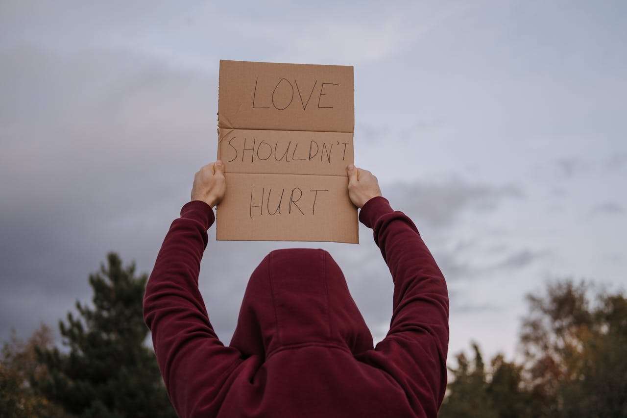 An anonymous person holds a cardboard sign reading 'Love Shouldn't Hurt' outdoors, advocating against violence.